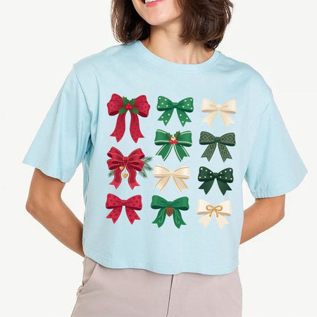 A cropped light blue t-shirt featuring a grid of twelve festive bow illustrations in red, green, and cream, worn by a model against a plain light background.