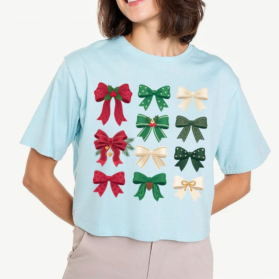 A cropped light blue t-shirt featuring a grid of twelve festive bow illustrations in red, green, and cream, worn by a model against a plain light background.