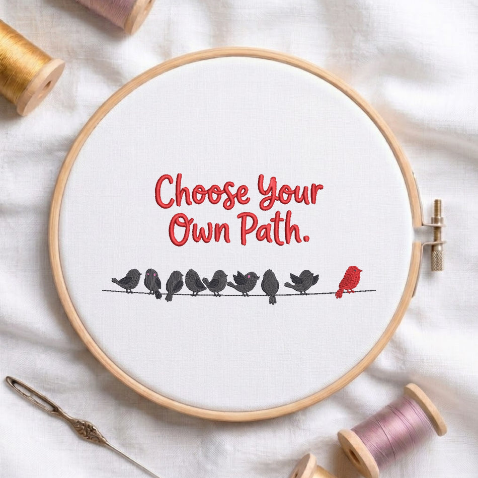 A white fabric stretched in a wooden embroidery hoop, featuring the red text "Choose Your Own Path." above a line of black birds sitting on a wire, with one distinct red bird standing apart at the end.