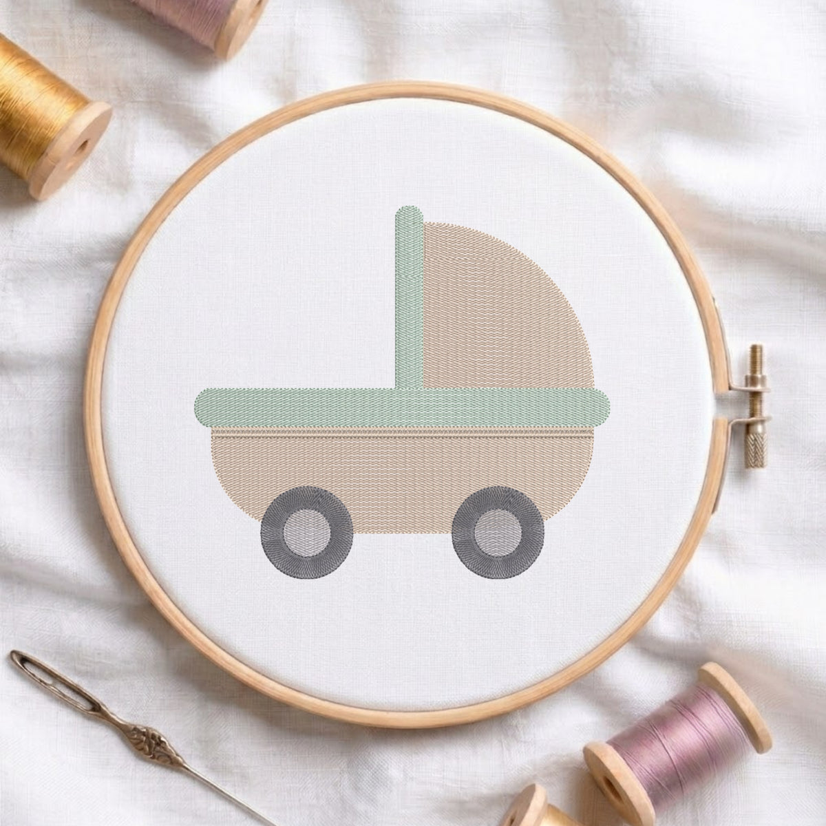 A round embroidery hoop holding white fabric with a simple baby stroller design in beige, sage green, and dark gray. A metal needle lies nearby, with spools of thread arranged around on a soft white fabric background.