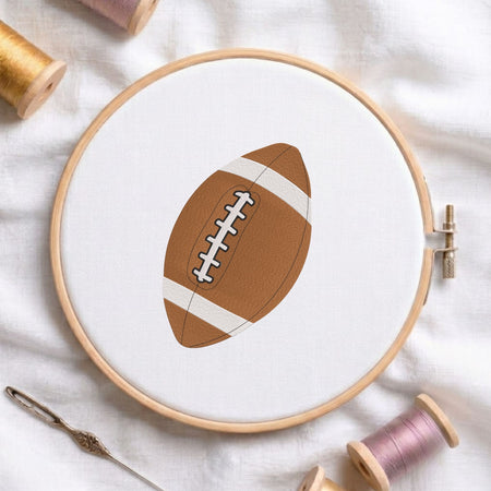 Football machine embroidery design displayed in a wooden embroidery hoop on white fabric with thread spools and a needle