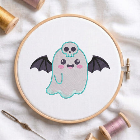 A circular embroidery hoop holding white fabric with a cute ghost design. The ghost has black bat wings, a small turquoise skull on its head, and pink cheeks.