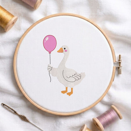 Cute duck wearing pink rain boots machine embroidery design displayed in a wooden hoop on white fabric with thread spools and a needle