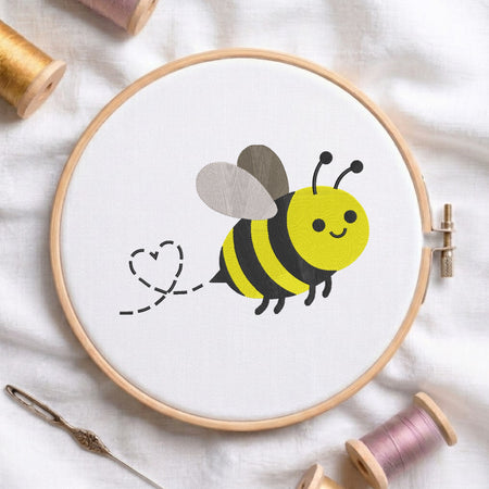 A wooden embroidery hoop holding white fabric with a cartoon bumblebee design in yellow, black, and gray. The hoop is styled on white fabric with spools of thread, a needle, and a thread spool visible around it.