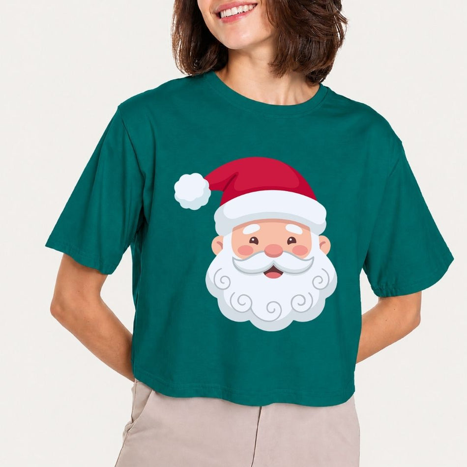A woman wearing a dark green short-sleeve crop top t-shirt with a cartoon Santa Claus face graphic on the front, paired with beige pants, against a light off-white background.