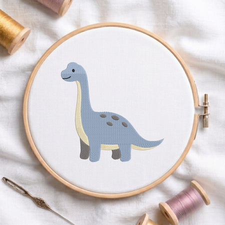 A circular embroidery hoop holding white fabric with a stitched long-neck dinosaur design in muted blue, cream, and gray. The hoop is styled on a white fabric surface with spools of thread and a needle around it.