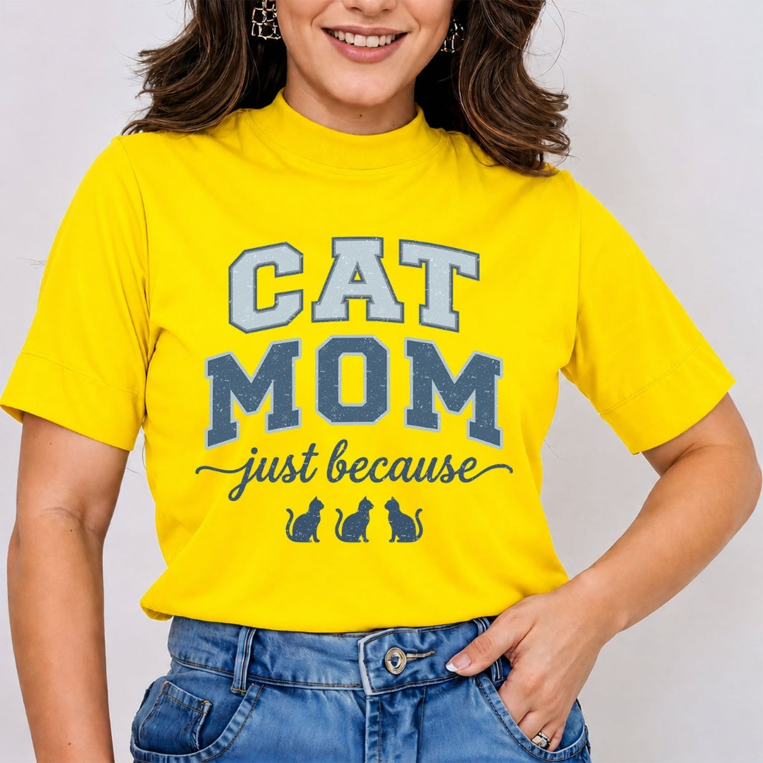Woman wearing a bright yellow mock neck t-shirt featuring a blue and white vintage-style "Cat Mom just because" graphic print with cat silhouettes, tucked into blue jeans.