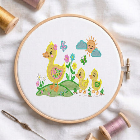 A circular embroidery hoop holding white fabric with a colorful stitched design of a mother duck, two chicks, flowers, butterflies, clouds, and a smiling sun. The hoop is styled on a white fabric background with spools of thread and a needle around it.