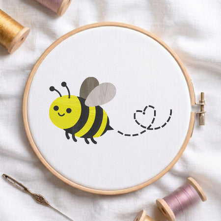 A circular embroidery hoop on white fabric, featuring a cartoon bee design in yellow, black, and gray with a dashed heart-shaped tail. Surrounding props include spools of thread, a needle, and a wooden stitch comb on a soft white fabric background.