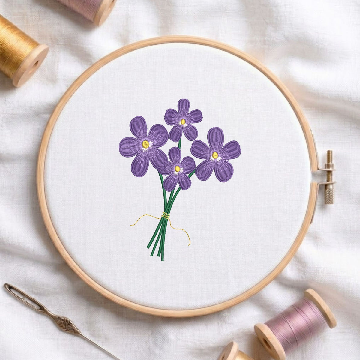 Purple flowers bouquet machine embroidery design displayed in a wooden hoop on white fabric with thread spools and a needle   