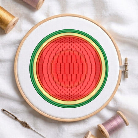 A circular embroidery hoop holding white fabric with concentric stitched rings in red, yellow, green, and white. Spools of thread and a needle are arranged around it on a white fabric background.