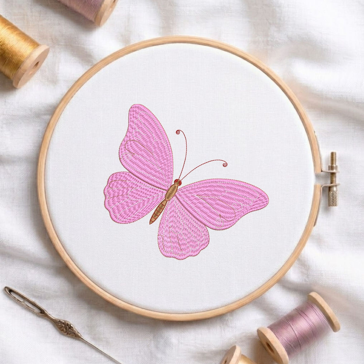 Elegant pink butterfly machine embroidery design displayed inside a round wooden hoop on white fabric with thread spools and a needle.