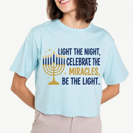 A woman wearing a light blue cropped t-shirt with a Hanukkah graphic that reads 'LIGHT THE NIGHT, CELEBRATE THE MIRACLES, BE THE LIGHT.' in navy and gold text alongside a gold menorah illustration.
