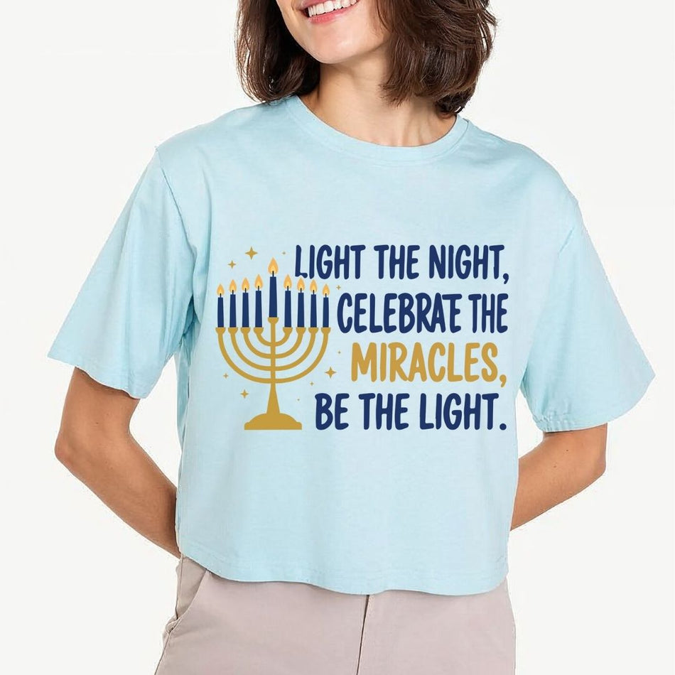A woman wearing a light blue cropped t-shirt with a Hanukkah graphic that reads 'LIGHT THE NIGHT, CELEBRATE THE MIRACLES, BE THE LIGHT.' in navy and gold text alongside a gold menorah illustration.
