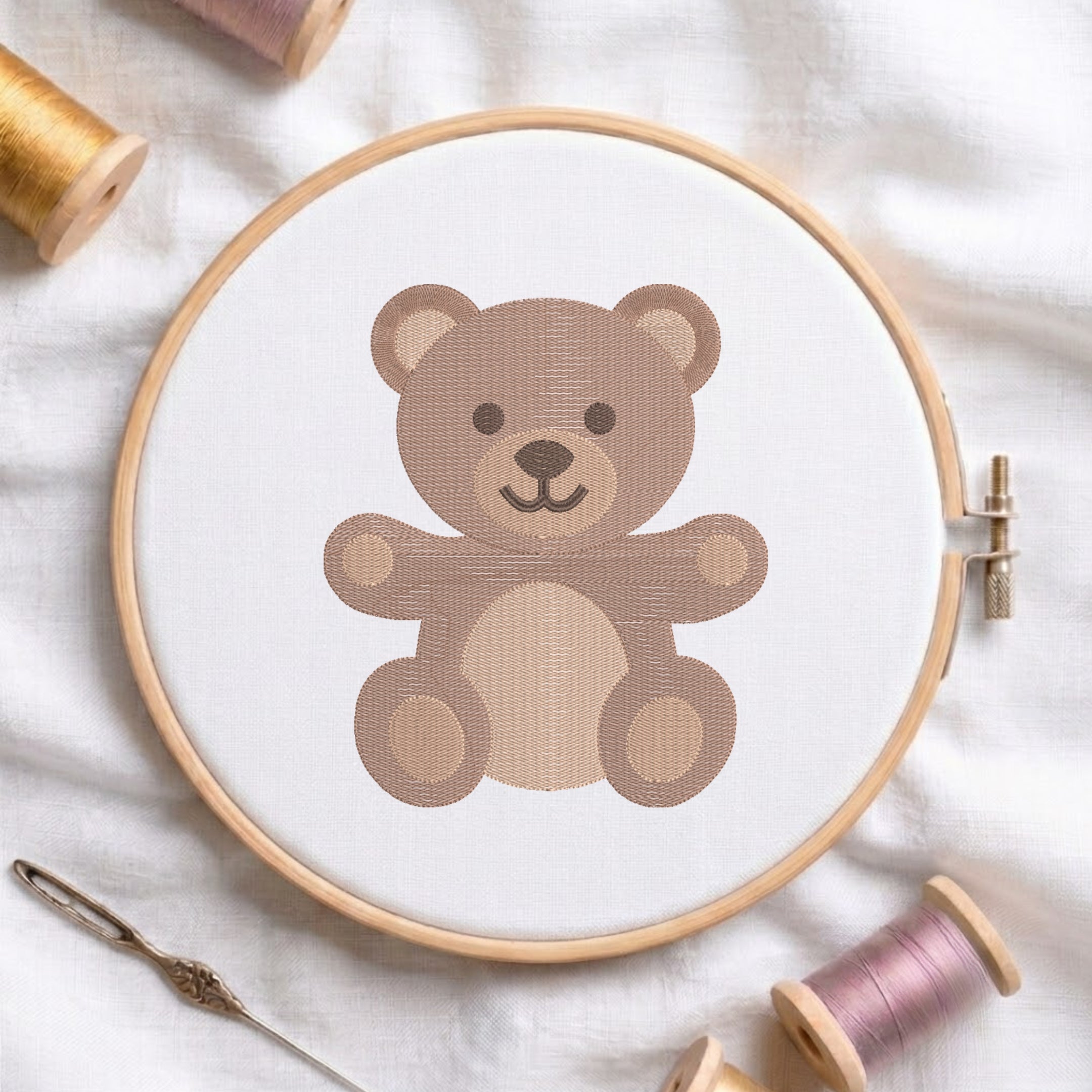 A wooden embroidery hoop holding white fabric with a stitched teddy bear design in shades of brown. A needle and spools of thread are arranged around it on a soft white fabric background.