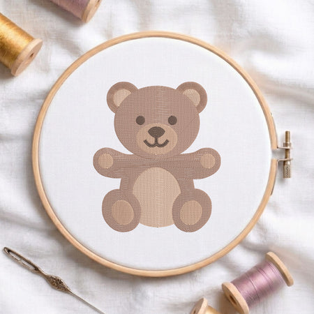 A wooden embroidery hoop holding white fabric with a stitched teddy bear design in shades of brown. A needle and spools of thread are arranged around it on a soft white fabric background.
