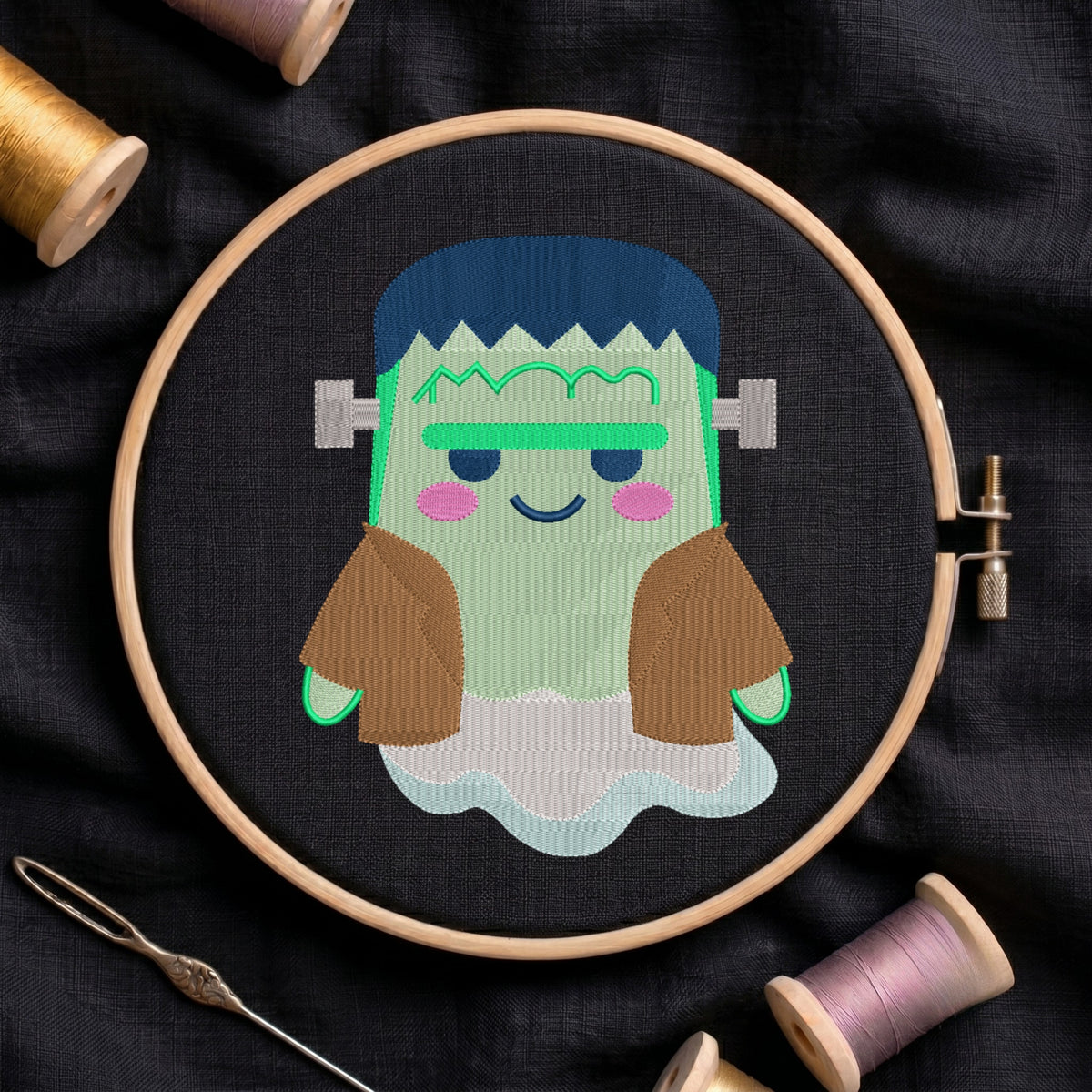 A circular embroidery hoop holds a stitched cartoon character resembling Frankenstein's monster, with blue hair, green skin, brown sleeves, and a white cloud-like base, set on dark fabric with thread spools and a needle around it.