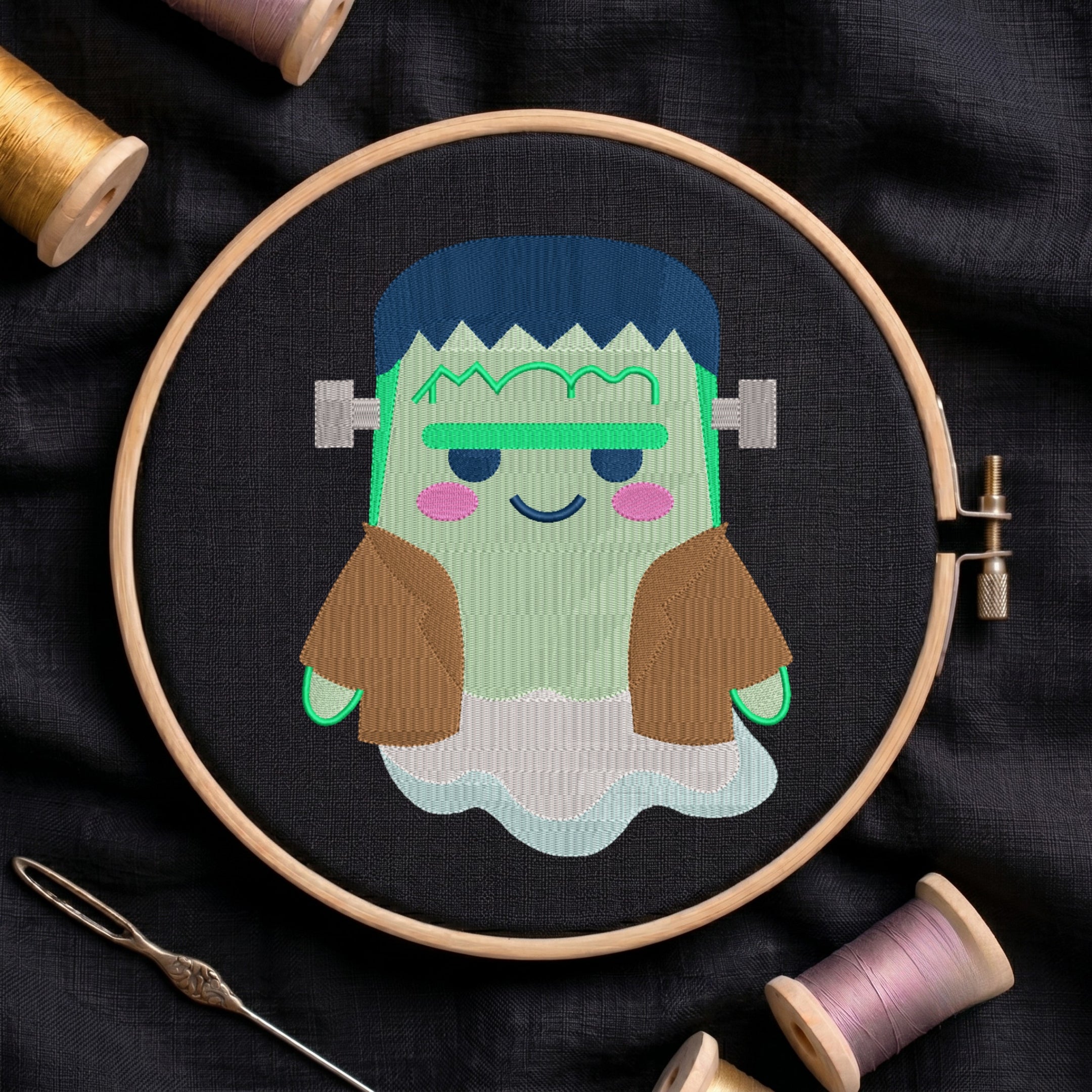 A circular embroidery hoop holds a stitched cartoon character resembling Frankenstein's monster, with blue hair, green skin, brown sleeves, and a white cloud-like base, set on dark fabric with thread spools and a needle around it.