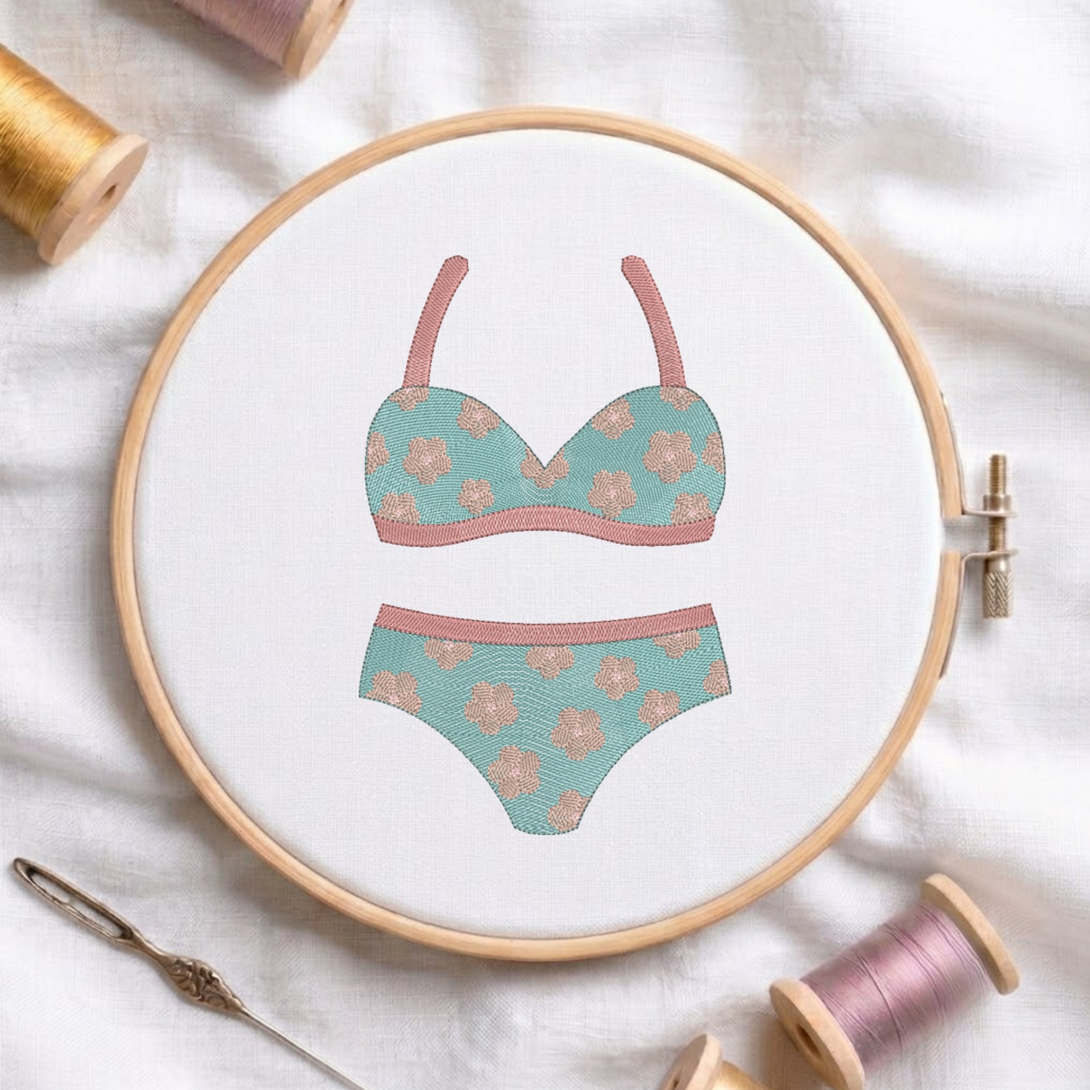 A circular embroidery hoop on white fabric displays a floral bikini design in mint green and dusty pink. Spools of thread, a needle, and soft fabric are arranged around the hoop in a styled flat-lay setup.