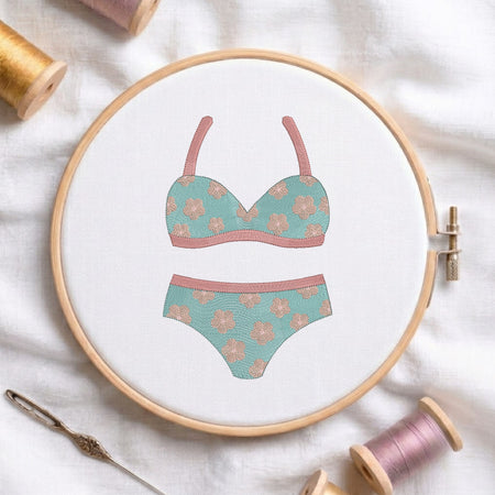 A circular embroidery hoop on white fabric displays a floral bikini design in mint green and dusty pink. Spools of thread, a needle, and soft fabric are arranged around the hoop in a styled flat-lay setup.