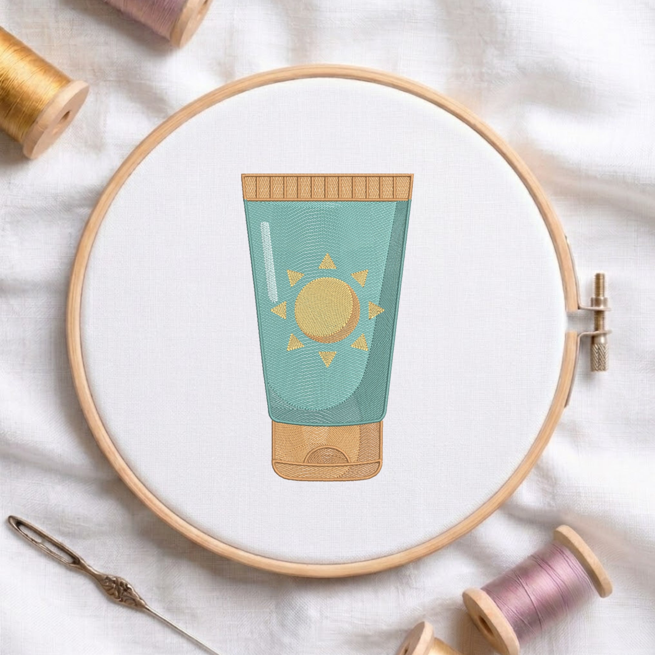 A circular embroidery hoop on white fabric holds a stylized embroidered tube illustration in teal and gold. Surrounding props include spools of thread and a needle on a soft white textile background.