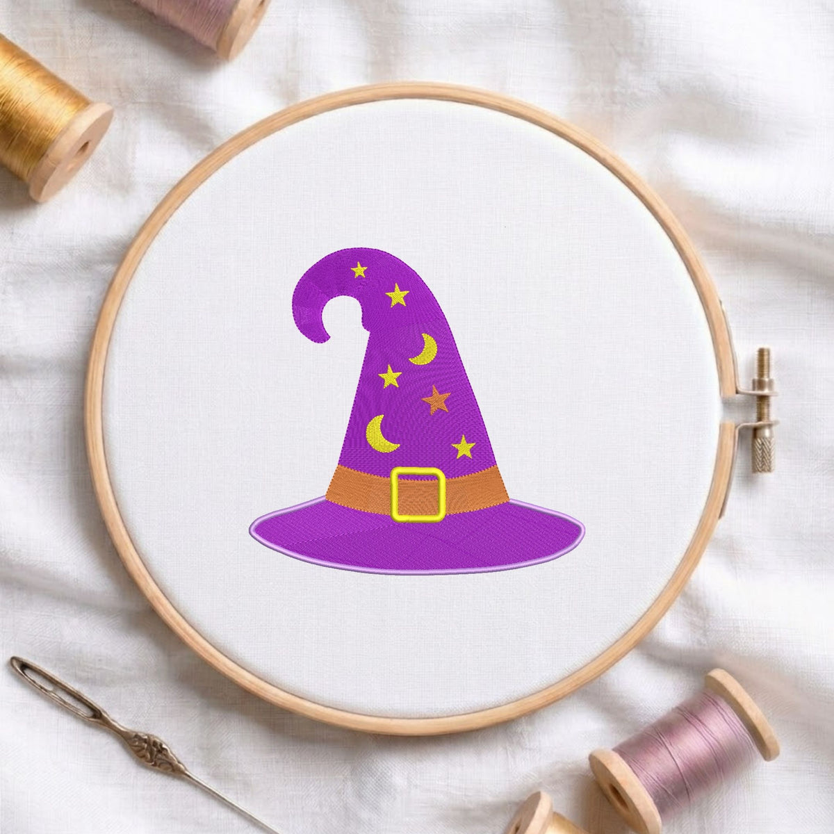 Purple witch hat machine embroidery design decorated with yellow stars and crescent moons, displayed inside a round wooden hoop on white linen fabric alongside thread spools and a needle.