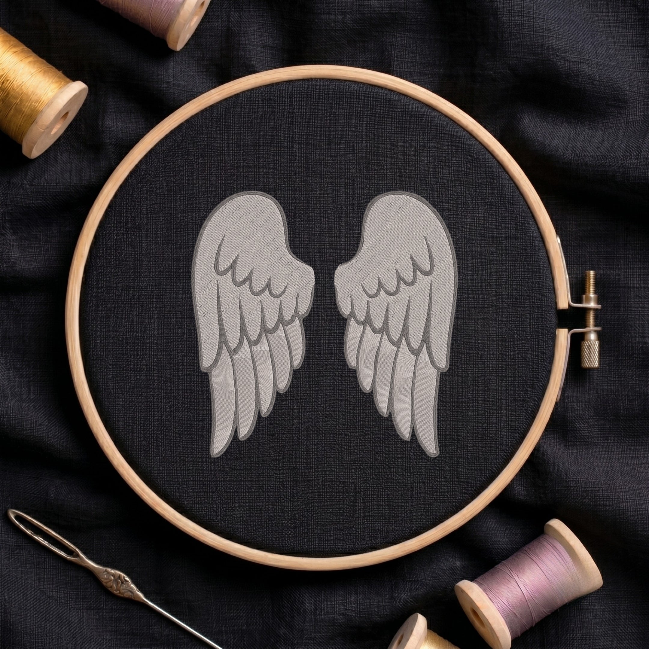 A wooden embroidery hoop holding black fabric with a gray embroidered pair of wings, surrounded by spools of thread and a needle on a dark fabric background.