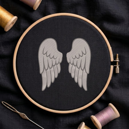 A wooden embroidery hoop holding black fabric with a gray embroidered pair of wings, surrounded by spools of thread and a needle on a dark fabric background.