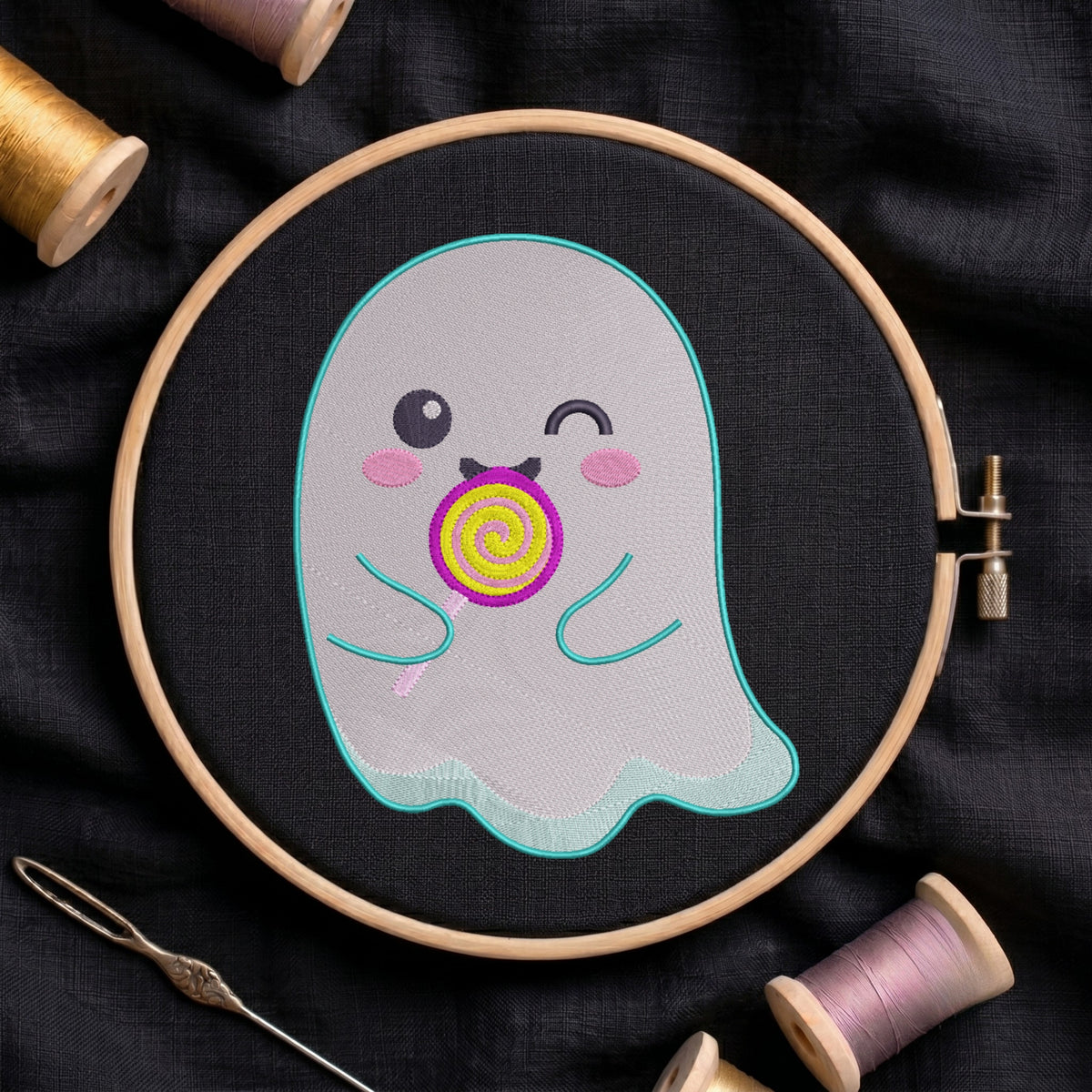 A close-up of a cute, winking ghost embroidery design featuring a colourful lollipop, stitched on black fabric inside a wooden embroidery hoop surrounded by spools of thread and a needle.