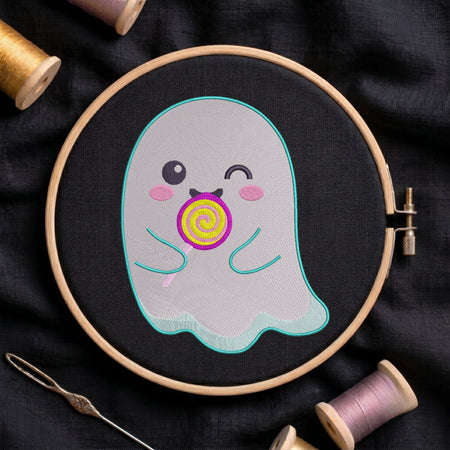 A close-up of a cute, winking ghost embroidery design featuring a colourful lollipop, stitched on black fabric inside a wooden embroidery hoop surrounded by spools of thread and a needle.