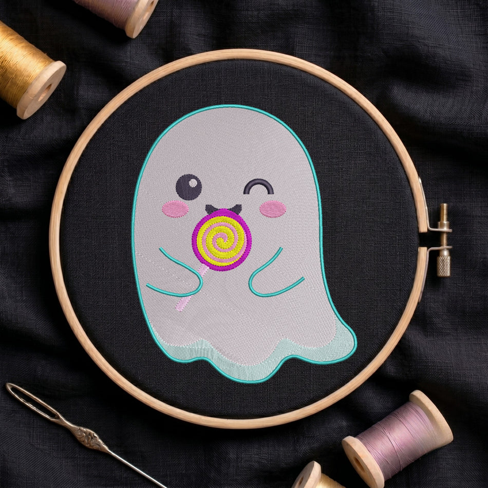 A close-up of a cute, winking ghost embroidery design featuring a colourful lollipop, stitched on black fabric inside a wooden embroidery hoop surrounded by spools of thread and a needle.