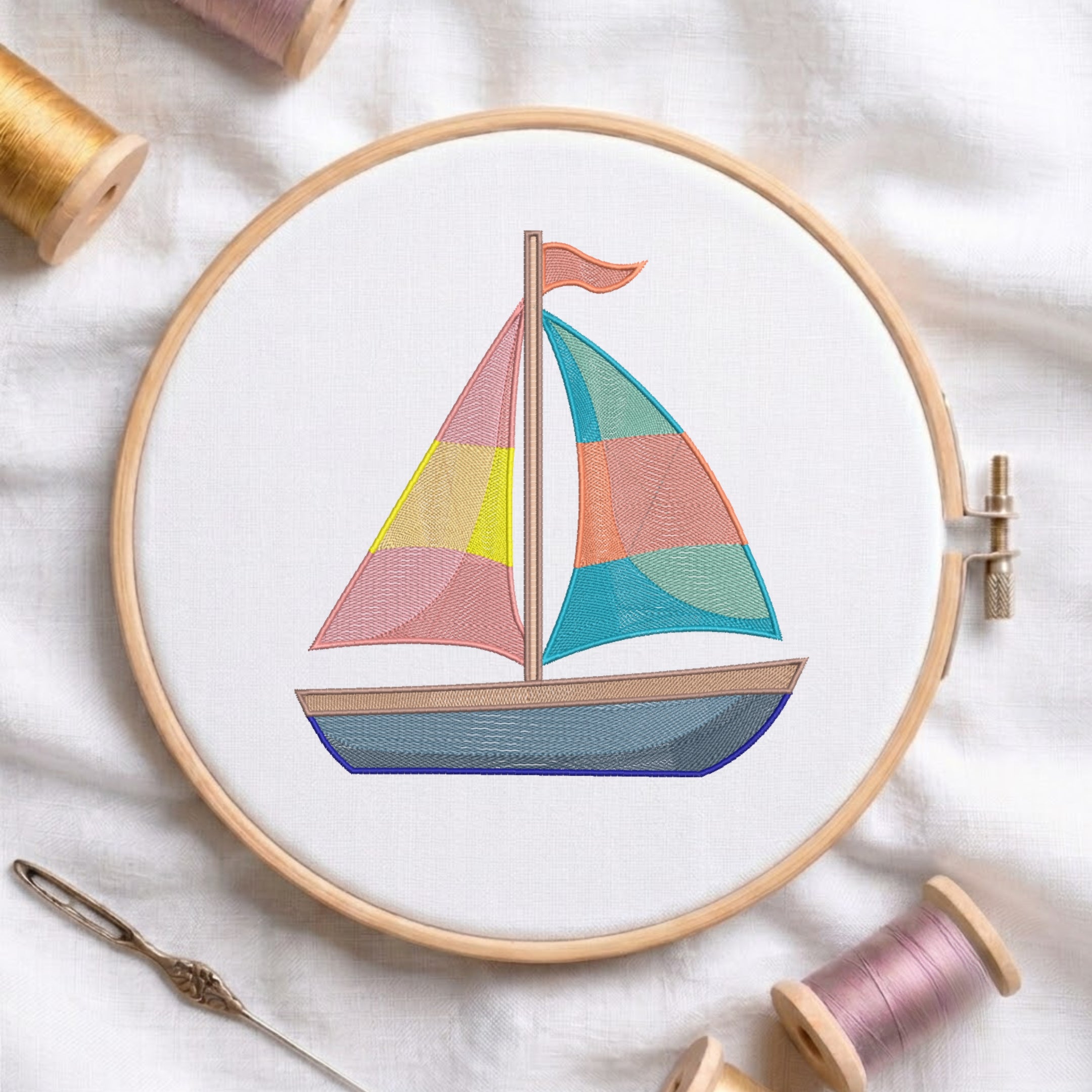 A wooden embroidery hoop holds a colorful sailboat design on white fabric, surrounded by spools of thread and a needle on a soft white textile background.