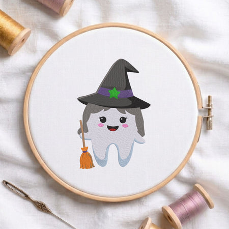 Cute witch tooth wearing a witch hat and holding a broomstick Halloween dental machine embroidery design shown in a wooden embroidery hoop on white fabric with thread spools and a needle