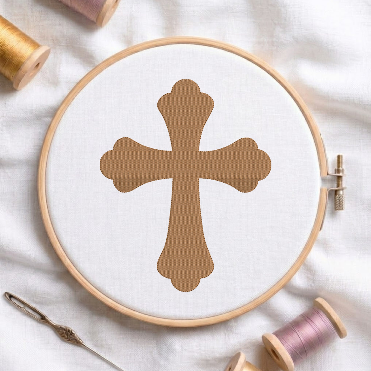 A wooden embroidery hoop holding white fabric with a stitched tan cross design, surrounded by spools of thread and a needle on a white fabric background.