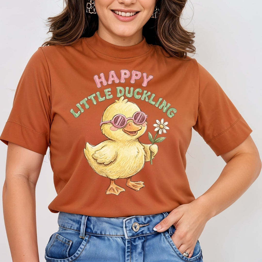 A woman wearing a rust-orange short-sleeve t-shirt with a cartoon graphic of a yellow duckling wearing pink sunglasses and holding a white flower, with the text 'HAPPY LITTLE DUCKLING' above it. She is also wearing blue jeans.