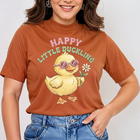 A woman wearing a rust-orange short-sleeve t-shirt with a cartoon graphic of a yellow duckling wearing pink sunglasses and holding a white flower, with the text 'HAPPY LITTLE DUCKLING' above it. She is also wearing blue jeans.