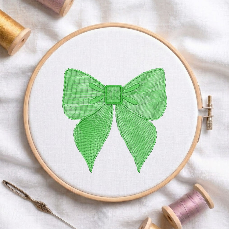 A wooden embroidery hoop holding white fabric with a green bow design stitched in the center, surrounded by spools of thread and a needle on a white fabric background.