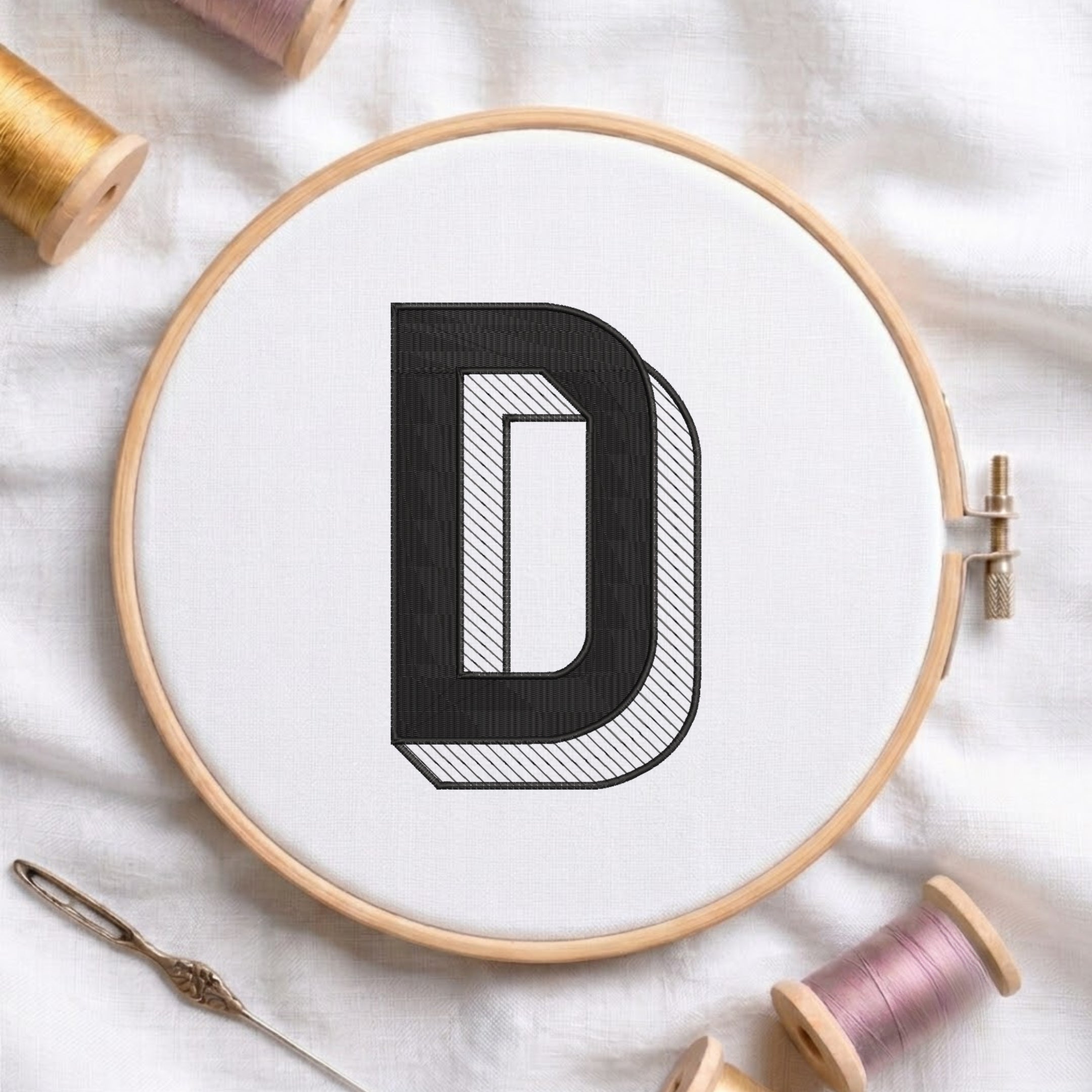 A wooden embroidery hoop holding white fabric with a large black letter D stitched in the center. A needle lies at the lower left, and spools of thread are arranged around the hoop on a white fabric background.