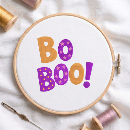 Orange and purple "Bo Boo!" Halloween lettering machine embroidery design featuring star and dot patterns, displayed inside a round wooden hoop on white fabric with thread spools and a needle.