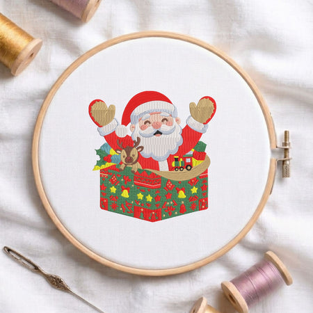 Joyful Santa Claus with a reindeer and toy train emerging from a festive patterned gift box machine embroidery design, displayed inside a round wooden hoop on white fabric with thread spools and a needle.