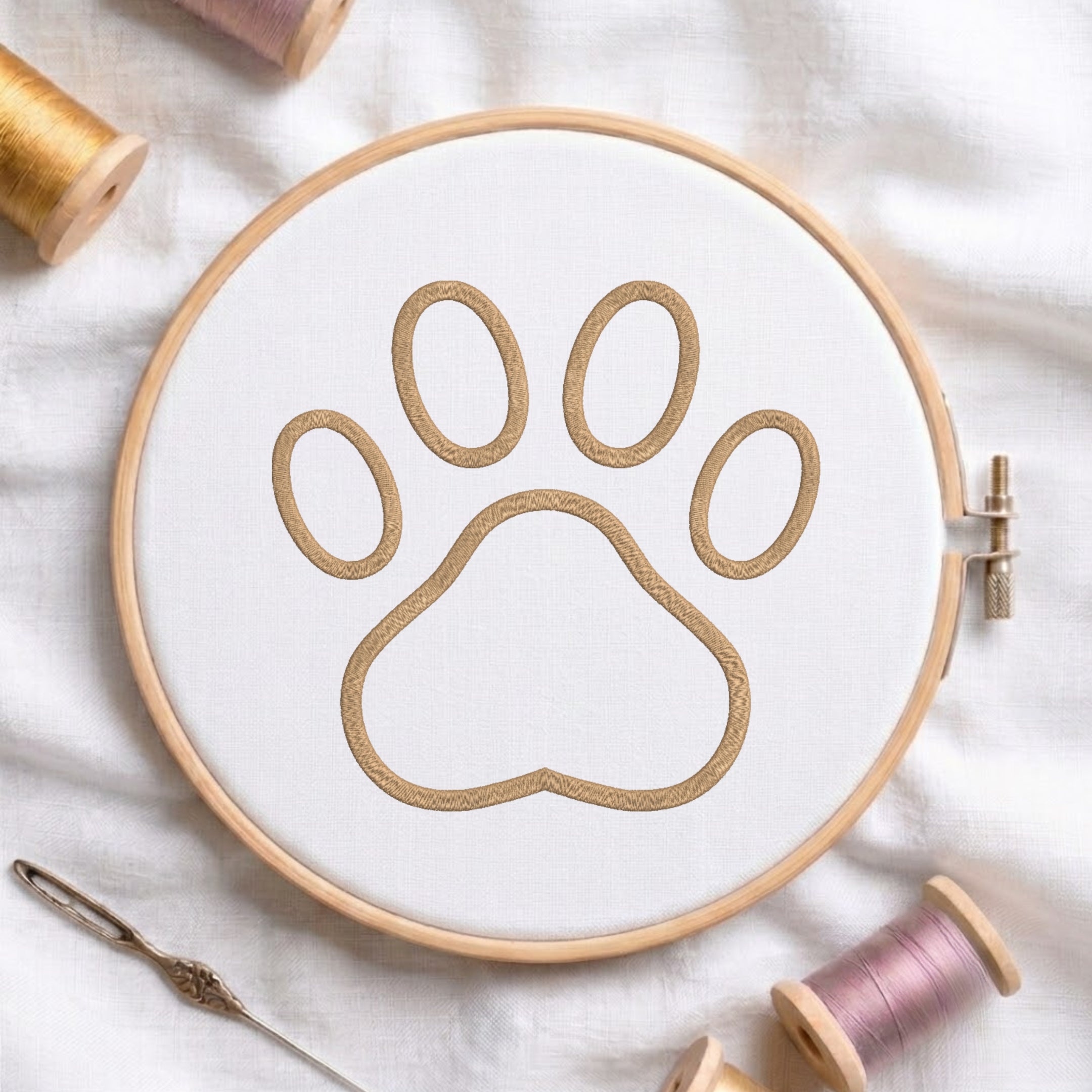 A circular embroidery hoop holding white fabric with a tan paw-print outline, surrounded by spools of thread and a needle on a white fabric background.