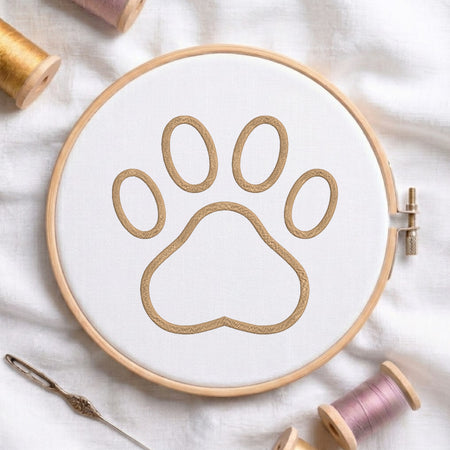 A circular embroidery hoop holding white fabric with a tan paw-print outline, surrounded by spools of thread and a needle on a white fabric background.