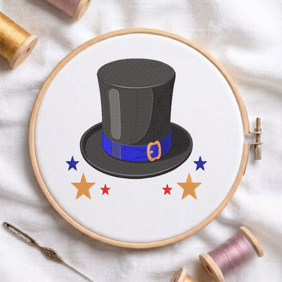 A circular embroidery hoop on white fabric holds a stitched design of a black top hat with a blue band and gold buckle, surrounded by blue and gold stars. A needle and spools of thread are arranged around it.