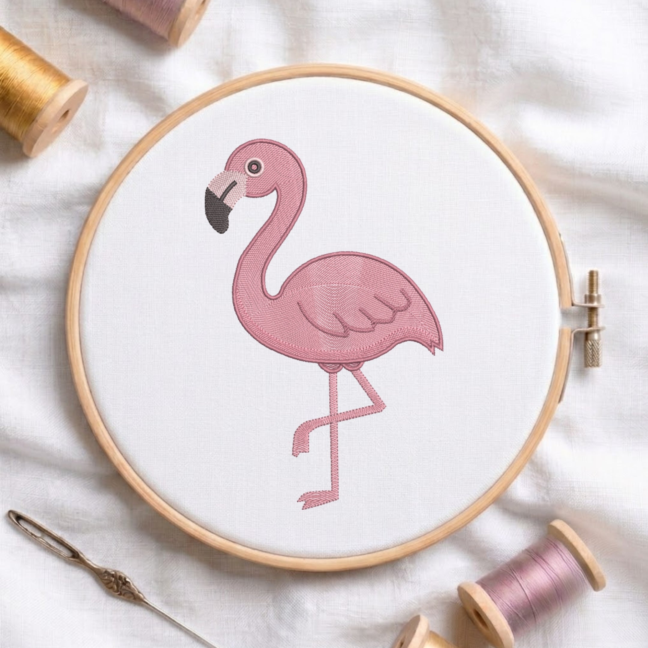 A pink flamingo embroidery design stitched on white fabric inside a wooden hoop, surrounded by spools of thread and a needle on a soft white fabric background.
