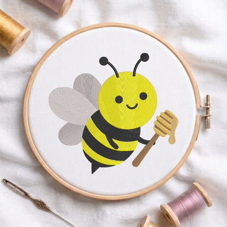 A circular embroidery hoop holding white fabric with a cartoon bee design in yellow, black, gray, and brown, surrounded by spools of thread and a needle on a white fabric background.
