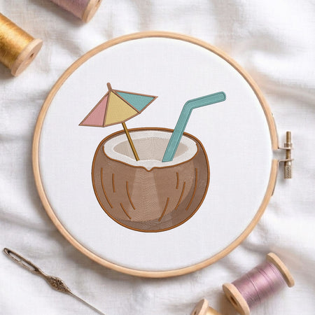 A circular embroidery hoop on white fabric, featuring a stitched design of a coconut drink with a small multicolor umbrella and teal straw. Spools of thread and a needle are arranged around the hoop.