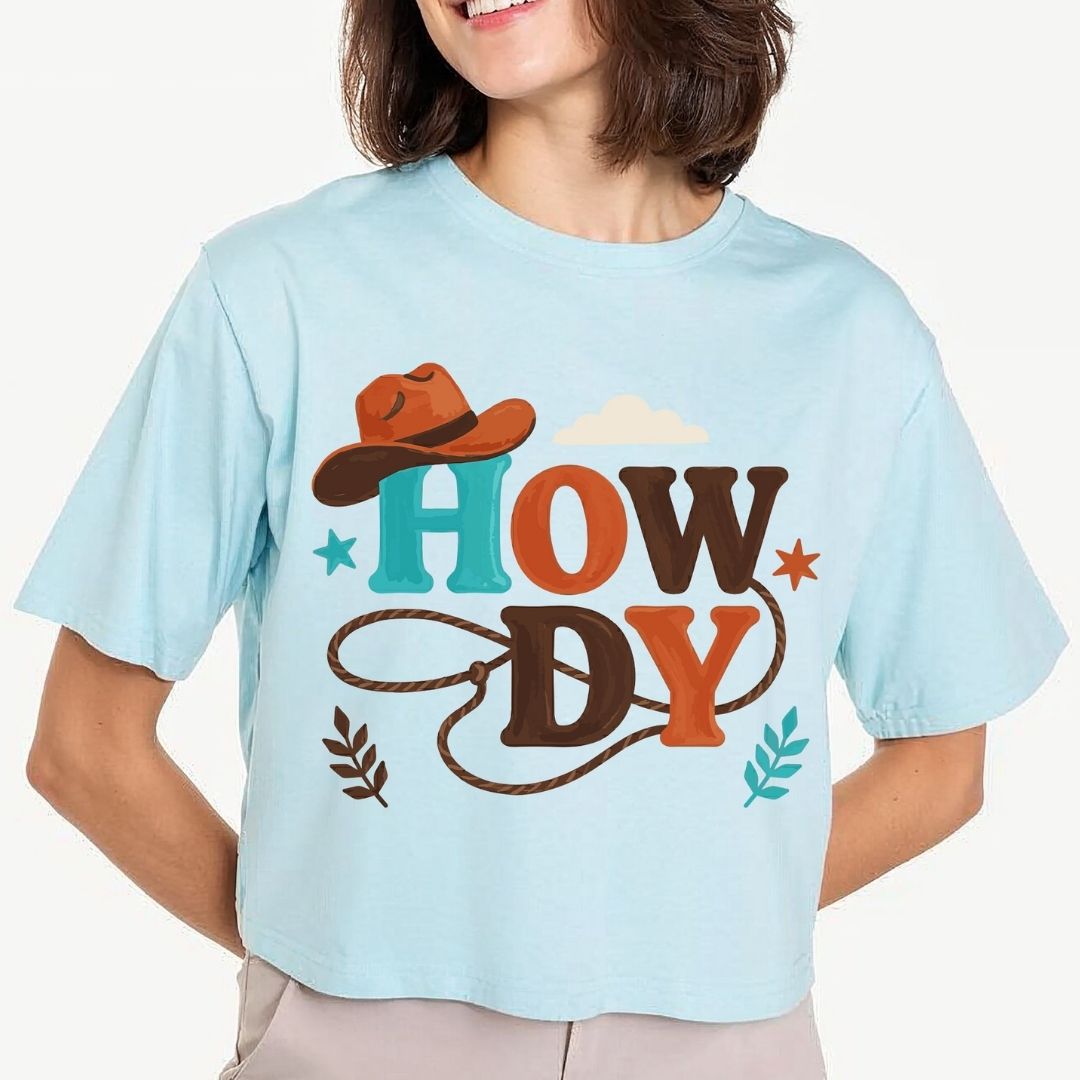 A woman modeling a light pastel blue crop top t-shirt featuring a large Western "Howdy" block letter design with a cowboy hat and lasso printed on the front.