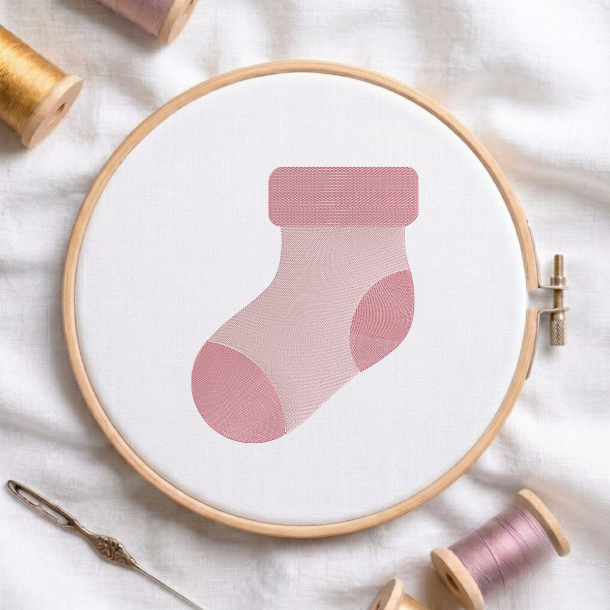 A pink sock-shaped embroidery design stitched inside a wooden hoop on white fabric, surrounded by spools of thread and a needle on a soft white background.