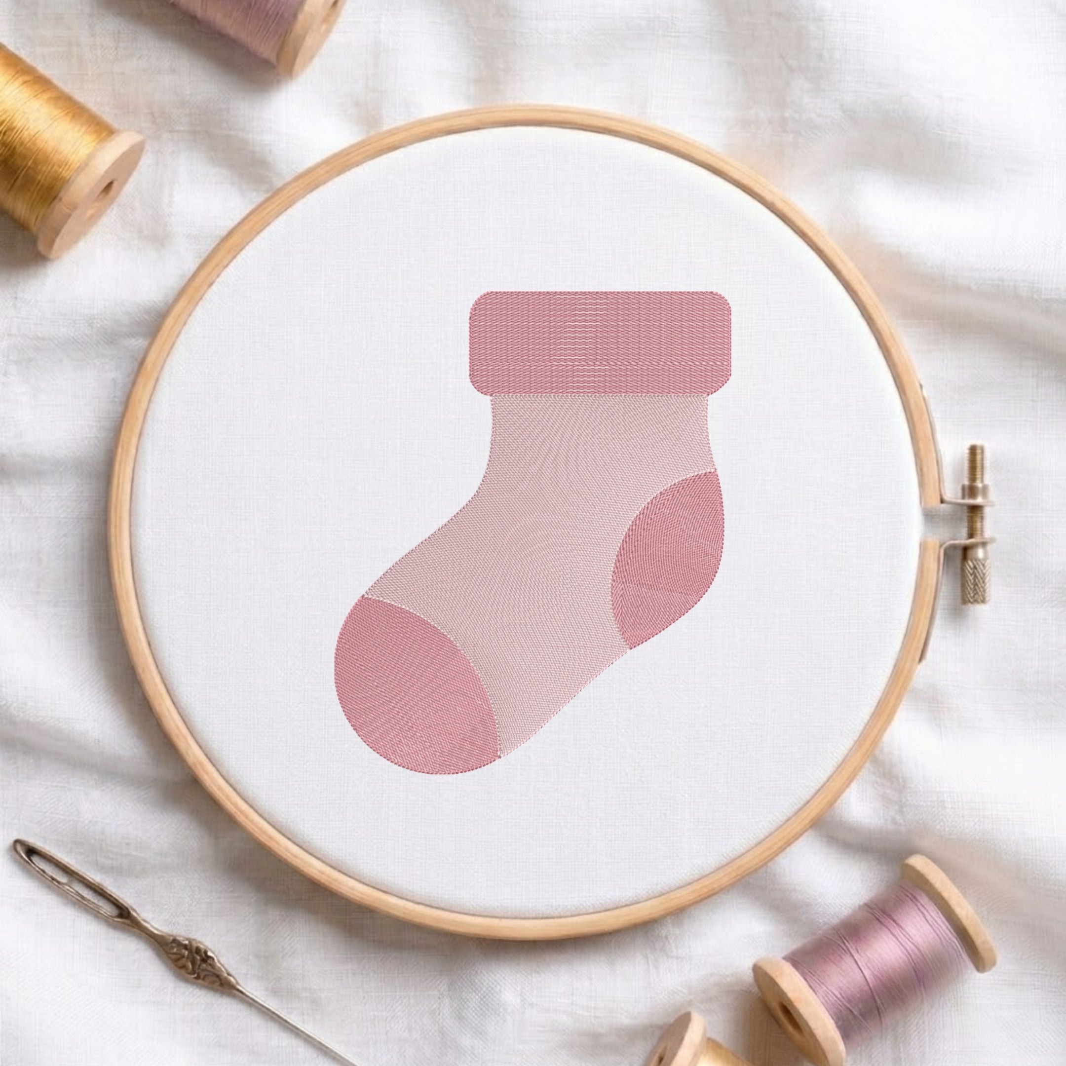 A pink sock-shaped embroidery design stitched inside a wooden hoop on white fabric, surrounded by spools of thread and a needle on a soft white background.