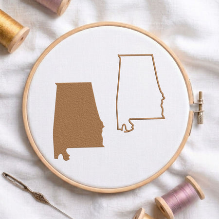 A wooden embroidery hoop on white fabric, showing two Alabama state outline designs in tan embroidery thread. A needle, additional thread spools, and soft fabric are arranged around it in a styled flat-lay.